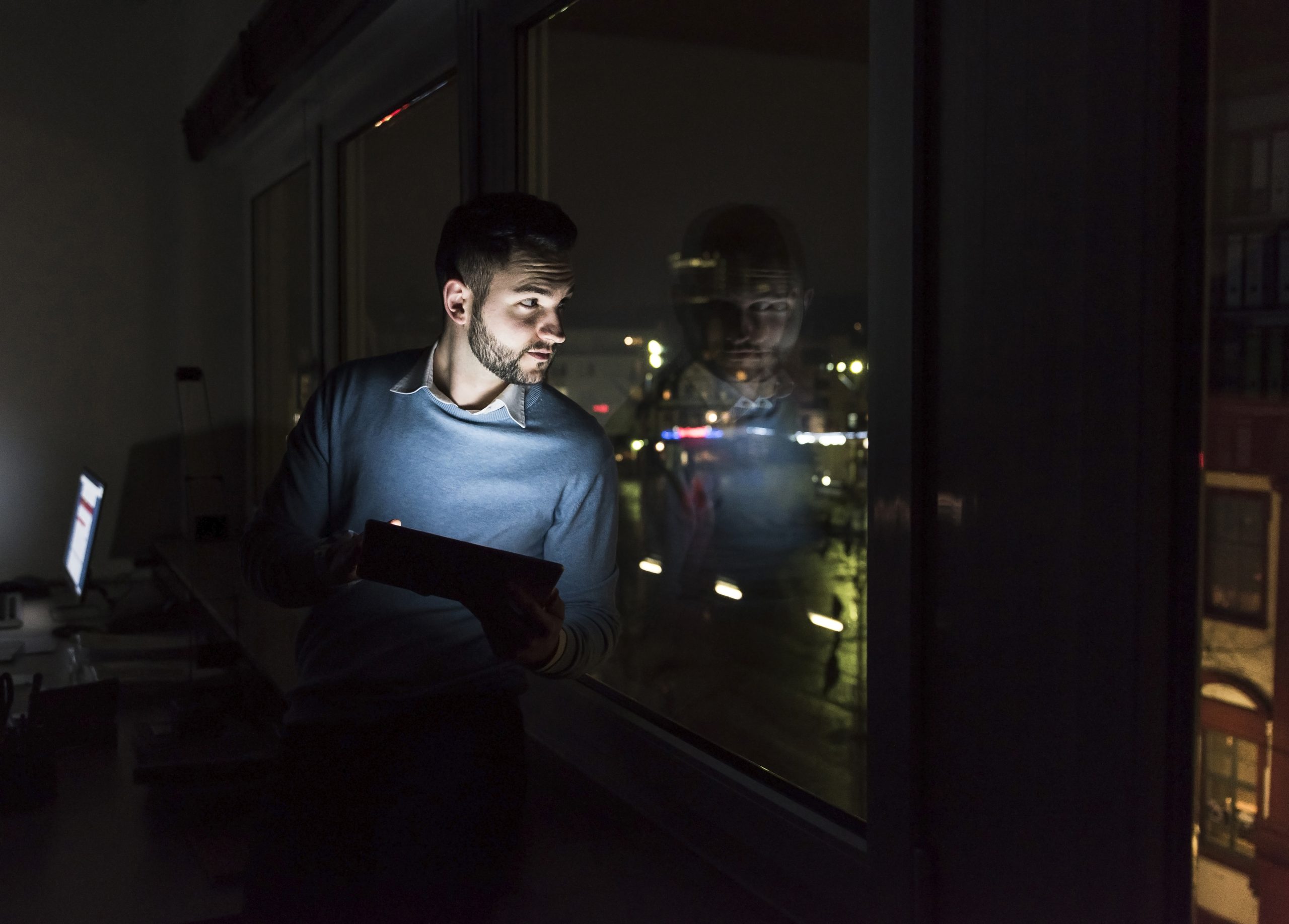 Businessman sitting with tablet on window sill in office at night looking out of the window